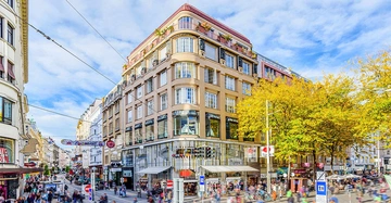 Menschen gehen an einem großen Gebäude mit vielen Fenstern vorbei in einer belebten Fußgängerzone mit Geschäften unter blauem Himmel und Herbstbäumen. Schilder: Turek BIPA Sports Direct.