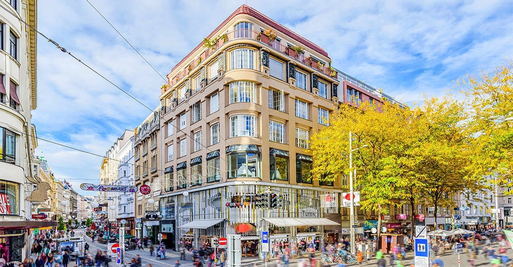 Menschen gehen an einem großen Gebäude mit vielen Fenstern vorbei in einer belebten Fußgängerzone mit Geschäften unter blauem Himmel und Herbstbäumen. Schilder: Turek BIPA Sports Direct.