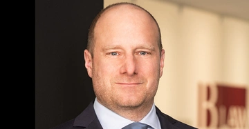 Mann im Anzug schaut ruhig nach vorne. Er steht in einem modernen Büro. Im Hintergrund sind verschwommene Bürowände und ein Schild mit dem Text BLaw erkennbar.