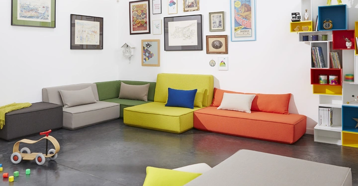 Mehrfarbige Sofas stehen in einer Ecke ein farbenfrohes Regal und verschiedene gerahmte Kunstwerke an weißen Wänden Ein Holzwagen und Spielzeugklötze liegen auf dem dunklen Boden.