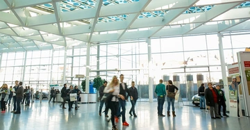 große, helle Messehalle mit Besuchern, Infoständen und Plakatständern. Lebendige Stimmung.