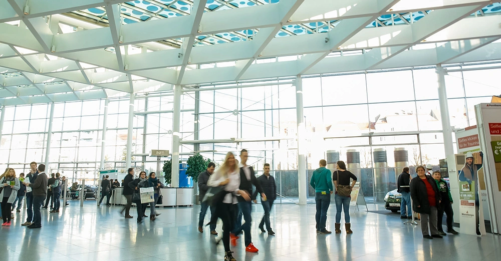 große, helle Messehalle mit Besuchern, Infoständen und Plakatständern. Lebendige Stimmung.