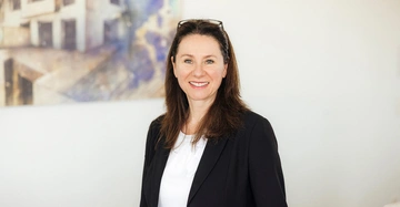 Frau lächelt Kamera an trägt schwarzes Sakko und weiße Bluse Hintergrund unscharfes Gemälde und weiße Wand.