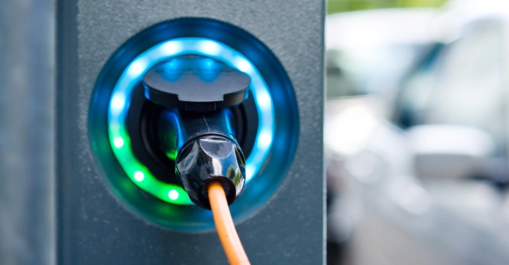 Ein schwarzer Ladestecker lädt ein Elektrofahrzeug an einer Ladesäule auf umgeben von einer blauen und grünen LED-Anzeige im Freien angrenzend an parkende Autos.