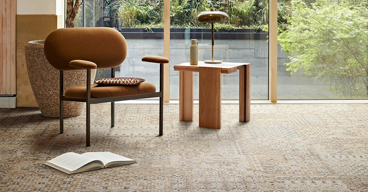 Brauner Sessel auf gemustertem Teppich neben einem Holztisch mit Lampe und Flasche in einem Raum mit Glaswand und Blick auf einen Garten. Ein aufgeschlagenes Buch liegt auf dem Boden.