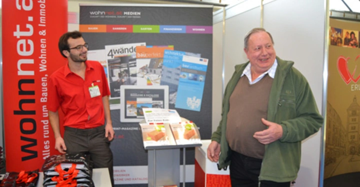 Galerie-Bild 9: Zwei Männer stehen an einem Messestand. Der links trägt ein rotes Hemd und steht hinter einem Tisch mit Broschüren. Der rechts trägt eine grüne Jacke und spricht. Umgebung Messe. Text wohnnet.at - Alles rund um Bauen, Wohnen & Immobilien.