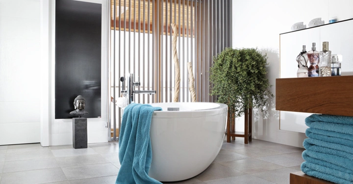 Galerie-Bild 6: Weiße ovale Badewanne mit blauer Handtuchablage steht vor vertikalen Holzlamellen im modernen Badezimmer ergänzt durch Buddha-Statue auf schwarzem Sockel einer Pflanze und hölzernen Regalen mit Kosmetikflaschen.