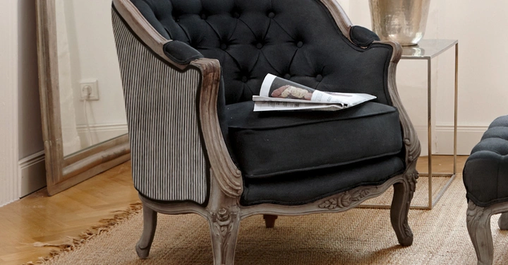 Galerie-Bild 2: Sessel mit schwarzer Polsterung und Holzrahmen hält eine Zeitung umgeben von einem Spiegel einem Metalltisch mit goldener Lampe und einem Teppich in einem hellen Raum.