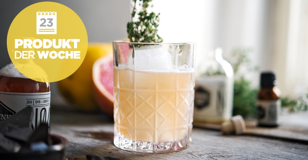 Ein Glas mit Getränk, Eiswürfel und Kräuterdekoration auf Holztisch; im Hintergrund eine Grapefruit und Getränkezutaten. Gelber Kreis mit der Aufschrift 23 PRODUKT DER WOCHE.