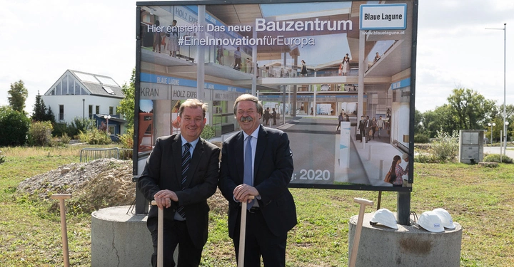 Zwei Männer halten Schaufeln vor einem Plakat mit der Aufschrift Hier entsteht Das neue Bauzentrum Eine Innovation für Europa Blaue Lagune Eröffnung 2020 Beide lächeln in einer offenen Wiesenlandschaft.