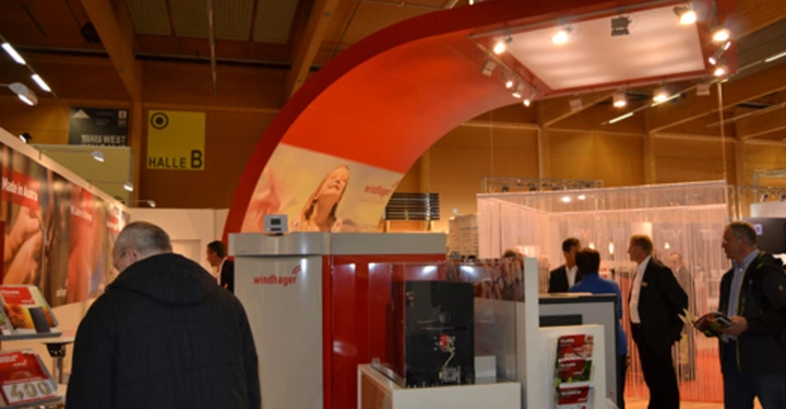 Galerie-Bild 14: Menschen besuchen den Windhager-Messestand in einer Halle während einer Messe. Der Stand zeigt Displays und Produkte, die Besucher begutachten. Schilder mit der Aufschrift "windhager" sind prominent sichtbar.