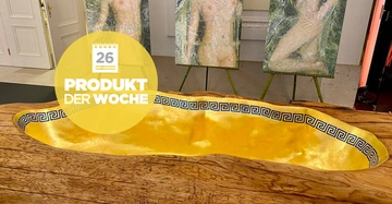 Gelbe Badewanne mit antikem griechischen Muster ruht auf hölzernem Rand in einem Ausstellungsraum zwei Bilder im Hintergrund. Text auf gelbem Kreis: 26 PRODUKT DER WOCHE.