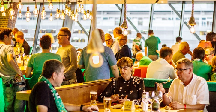 Galerie-Bild 2: Menschen unterhalten sich und trinken in einem gut beleuchteten Restaurant oder einer Bar mit großen Fenstern und modernen Hängelampen im Hintergrund.