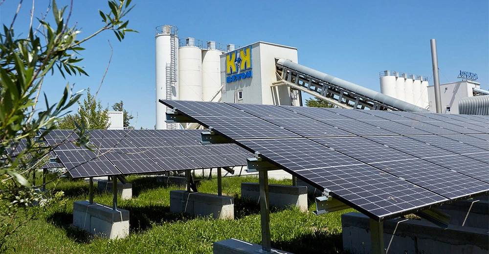 Solarmodule werden in der Nähe eines Betonwerks mit mehreren Silos betrieben umgeben von Grünflächen bei sonnigem Wetter. Text auf dem Gebäude: "K K BETON".