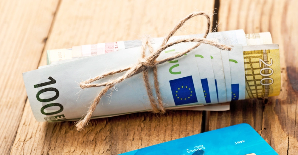 Ein Bündel Euro-Geldscheine wird mit einem Bindfaden zusammengehalten, neben einer blauen Kreditkarte, auf einem Holztisch liegend.