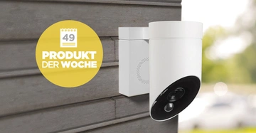 Weiße Überwachungskamera, an einer grauen Holzplatte montiert. Gelber Kreis mit der Aufschrift Woche 49 Produkt der Woche. Unscharfer, grüner Hintergrund.