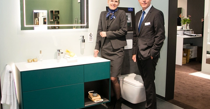 Galerie-Bild 2: Zwei Personen posieren in einem modernen Badezimmerdesign-Ausstellungsraum eines Bademöbelunternehmens mit grünem Waschtisch, großem Spiegel und Handtuch auf einem Handtuchhalter darunter
