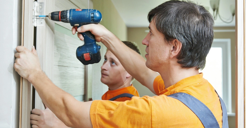 Arbeiter im Blaumann mit Akkuschrauber zieht Schrauben am Türbeschlag fest. Zweiter Arbeiter im Hintergrund hält das Türblatt fest. 