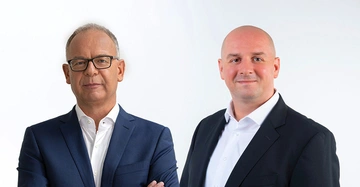 Zwei Männer tragen Anzüge und weiße Hemden posieren nebeneinander vor einem weißen Hintergrund der Mann links mit Brille hat die Arme verschränkt der Mann rechts lächelt leicht