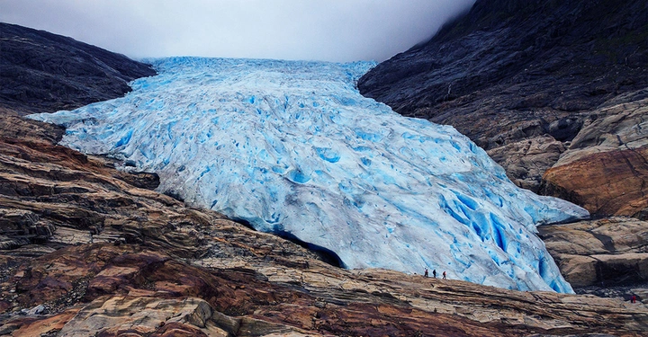 Blauer Gletscher erstreckt sich zwischen felsigen Bergen wobei im Vordergrund kleine Menschen auf dem Gestein stehen und aufsteigen während der Himmel grau und bewölkt ist