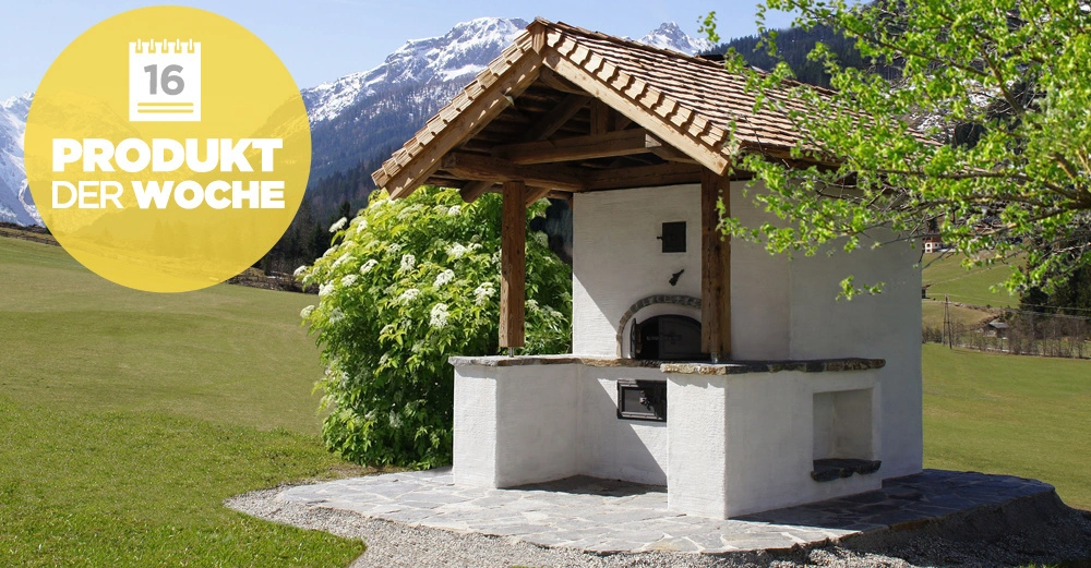 Ein gemauerter Außenofen steht unter einem hölzernen Dach auf einer mit Steinen gepflasterten Fläche in einer Berglandschaft mit grünen Wiesen und blühenden Bäumen. Text: 16 PRODUKT DER WOCHE.