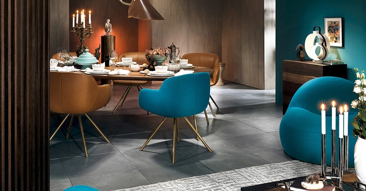 Essgruppe aus blauen und braunen Stühlen umgibt einen gedeckten runden Tisch in einem modern eingerichteten Raum mit Kerzenständern und Dekor; beleuchtete Stehlampe und Kommode im Hintergrund.