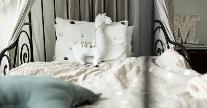 Galerie-Bild 2: Ein weißes Stofftier Lama sitzt auf einem Bett mit beigefarbener Decke und weißen Kissen im Hintergrund in einem gemütlichen, hellen Schlafzimmer.