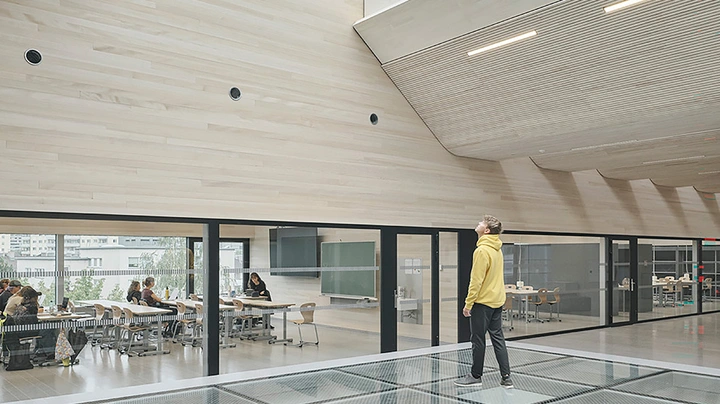 Galerie-Bild 2: Junger Mann in gelbem Hoodie steht vor Klassenzimmer mit verglaster Seitenwand und blickt in Holzdachverbau.
