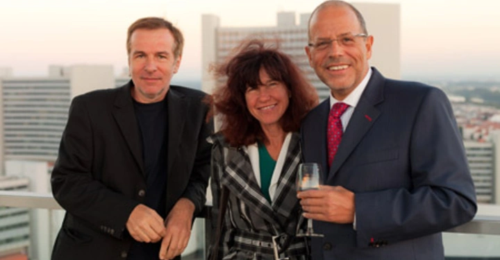 Galerie-Bild 37: Drei Personen stehen auf einem Balkon Ein Mann lehnt sich an das Geländer Eine Frau steht in der Mitte Ein anderer Mann im Anzug hält ein Glas Stadtlandschaft im Hintergrund