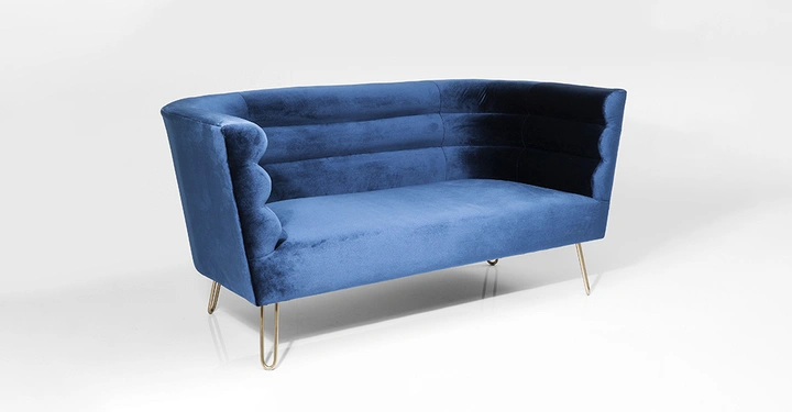 Blaues Sofa mit samtartiger Polsterung sitzt auf dünnen goldenen Beinen im leerer weißer Raum