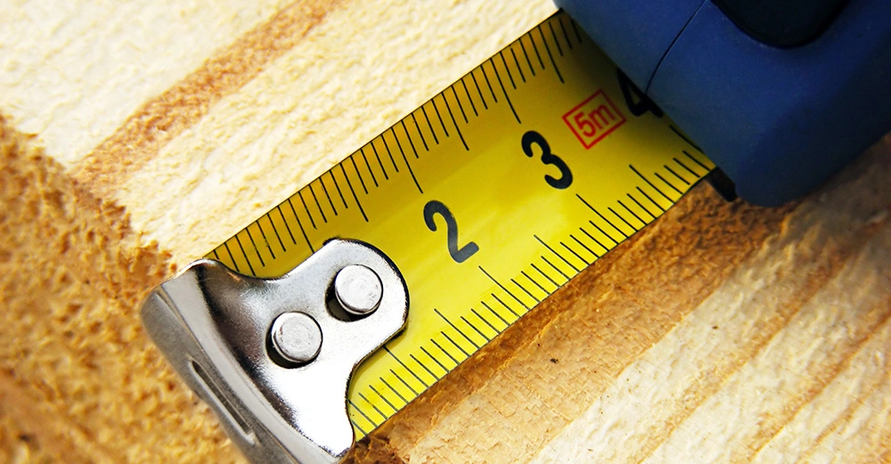 Ein gelbes Maßband mit schwarzen Zahlen und Markierungen und der roten Zahl 5m wird über ein Holzstück gespannt