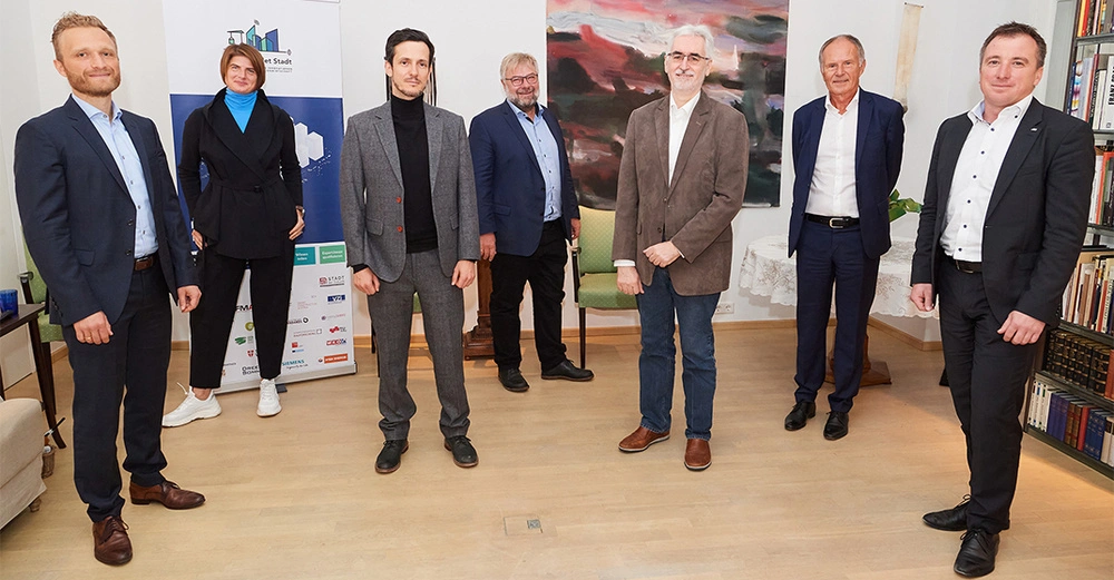 Sieben Personen stehen in einem Raum, tragen Businesskleidung und lächeln in die Kamera. Im Hintergrund befinden sich ein Poster mit Logos und ein abstraktes Gemälde. Links sind Bücherregale.