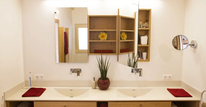 Galerie-Bild 7: Doppelwaschbecken mit roten Handtüchern, Holzschränken und zwei Spiegeln; im Hintergrund Fenster und Handtuchhalter; in der Mitte steht ein dekorativer Pflanzenbehälter auf der Ablagefläche.