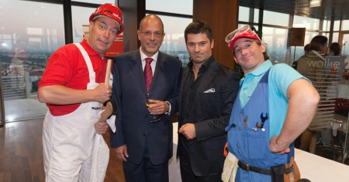 Galerie-Bild 49: Vier Männer stehen in einem eleganten Raum mit großen Fenstern. Zwei tragen Anzüge und die anderen zwei sind in Handwerkerkleidung. Einer hält ein Glas Getränk.