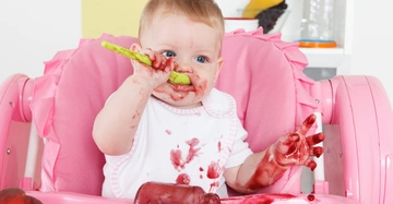 Baby isst mit einem grünen Löffel Beikost im rosa Hochstuhl und ist mit Essen verschmiert im Hintergrund Obst und Regale.