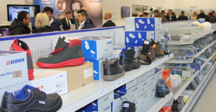 Galerie-Bild 3: Regale mit Sicherheitsschuhen stehen in einem gut beleuchteten Geschäft Ladenpersonal und Kunden in Geschäftskleidung unterhalten sich im Hintergrund Text: BERNER Berner Safety Shoes