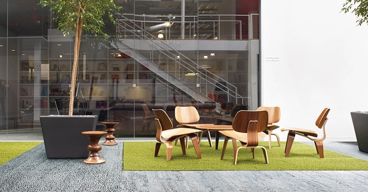 Galerie-Bild 3: Holzmöbel stehen zusammen auf einem Teppich auf grünem Rasen in einem modernen Büro mit einem hellen Raum im Hintergrund durch eine Glaswand und eine Treppe sichtbar