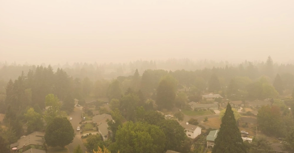 Luftaufnahme einer Wohnsiedlung, die in dichten Rauch gehüllt ist, vermutlich verursacht durch Waldbrände. Die Sicht ist stark eingeschränkt, die Landschaft wirkt vernebelt und orange-gelblich getönt. Bäume und Häuser sind nur schemenhaft erkennbar.