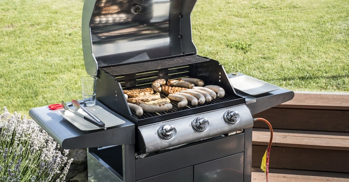 Grill mit Würstchen und Fleisch grillend im Garten. Links sind Grillutensilien und ein Glas auf einem Tablett. Im Hintergrund Rasen und Holzterrasse.