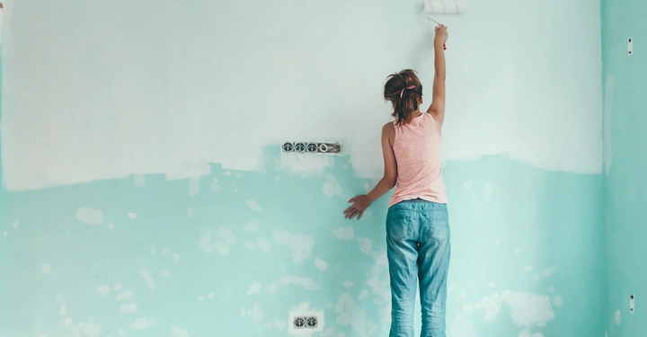 Person streicht eine Wand mit einer Farbrolle in einem Raum im Innenbereich die Wand ist teilweise mintgrün und teilweise weiß die Person trägt ein gestreiftes Oberteil und blaue Jeans
