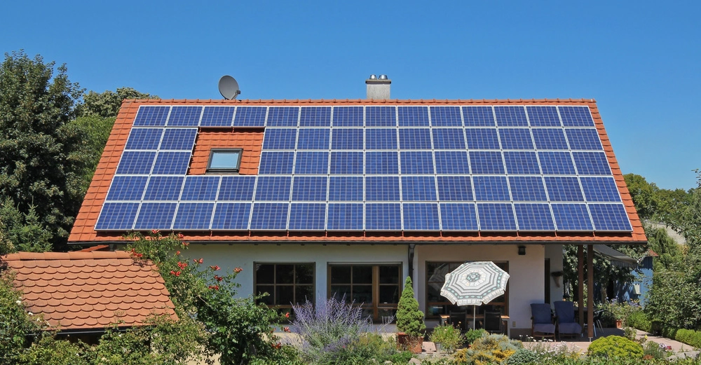 Haus mit Solarpaneelen bedeckt das Dach in einem üppig grünen Garten während blauer Himmel den sonnigen Tag unterstreicht.