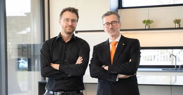 Tobias Hüttl und Georg Niedersüß stehen mit verschränkten Armen nebeneinander in der Küche vom Floating House. Hüttl leger im scharzen Hemd, Niedersüß mit Anzug und oranger Krawatte.