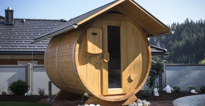 Galerie-Bild 3: Holzsauna in Form eines Fasses steht im Garten neben einem modernen Haus in einer bergigen Gegend mit Bäumen im Hintergrund.