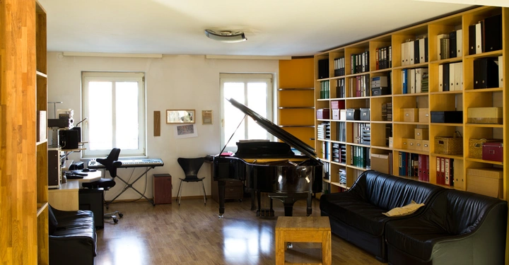 Galerie-Bild 2: Großes Zimmer mit einem Flügel und einem Keyboard in der Mitte mehrere Regale voller Akten und Schachteln an der rechten Wand eine schwarze Ledercouch und ein Schreibtischstuhl.