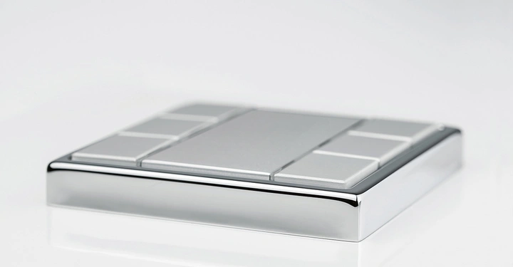 Silberner rechteckiger Gegenstand mit mehreren grauen rechteckigen Tasten in verschiedenen Größen auf der Oberseite liegt auf einer glänzenden weißen Oberfläche in einem minimalistischen Umfeld.