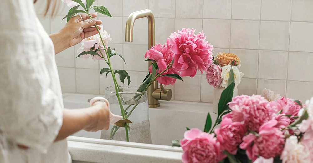 Frau arrangiert Pfingstrosen in Vase am Spülbecken mit Messingarmatur. Nachhaltig dekorieren mit frischen Blumen in moderner Küche.