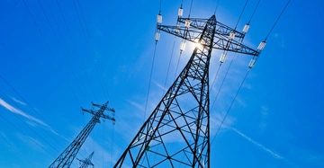 Strommast steht still vor einem klaren blauen Himmel mit Sonne im Hintergrund mehrere Drähte erstrecken sich seitlich von ihm.