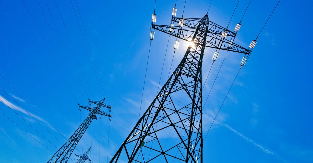 Strommast steht still vor einem klaren blauen Himmel mit Sonne im Hintergrund mehrere Drähte erstrecken sich seitlich von ihm.