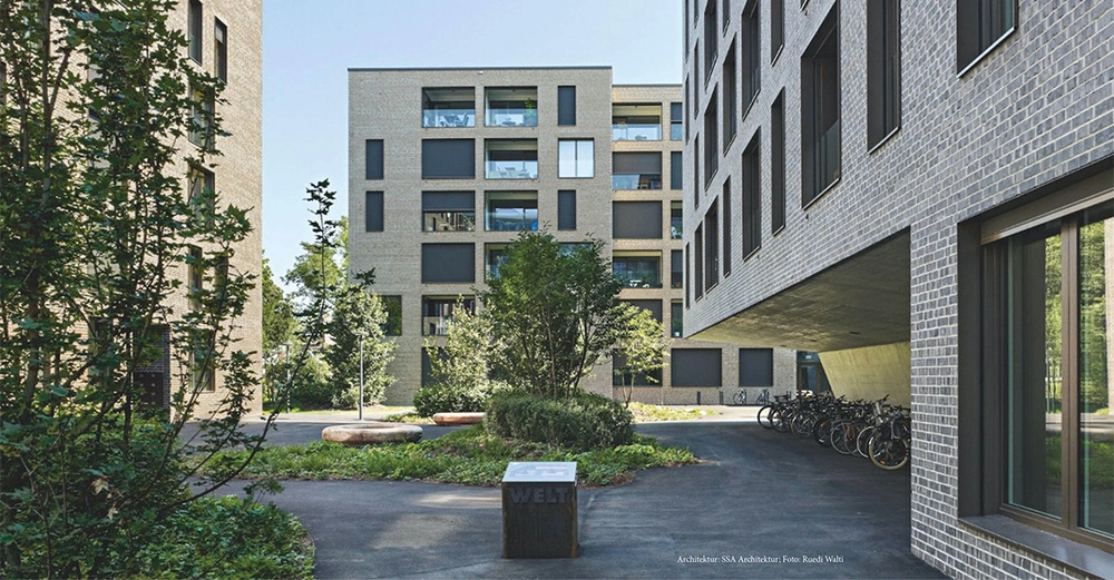 Mehrstöckige moderne Gebäude aus Backstein umgeben einen begrünten Hof mit Radständern und Sitzbänken. Text auf einem Steinwürfel: "WELT". Text unten rechts: "Architektur SSA Architekten Foto Ruedi Walti".