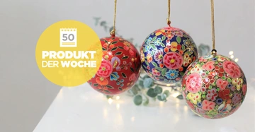 Drei weihnachtliche Kugeln mit Blumenmustern hängen an goldenen Fäden; links ein gelber Kreis mit Text 50 PRODUKT DER WOCHE; Hintergrund unscharf, weiße Oberfläche mit kleinen Lichtern und Grün.